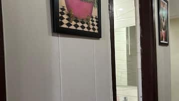 A hallway with a framed picture of a pink bathtub on a checkered floor, a white wall, and a door leading to a bathroom.