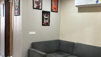 A corner room with a grey sofa, a wall-mounted air conditioner, and framed artwork.