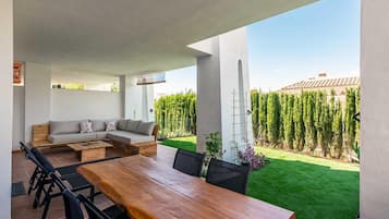 Een terras buiten met een houten tafel en stoelen, een bank, en uitzicht op een verzorgd gazon en bomen.