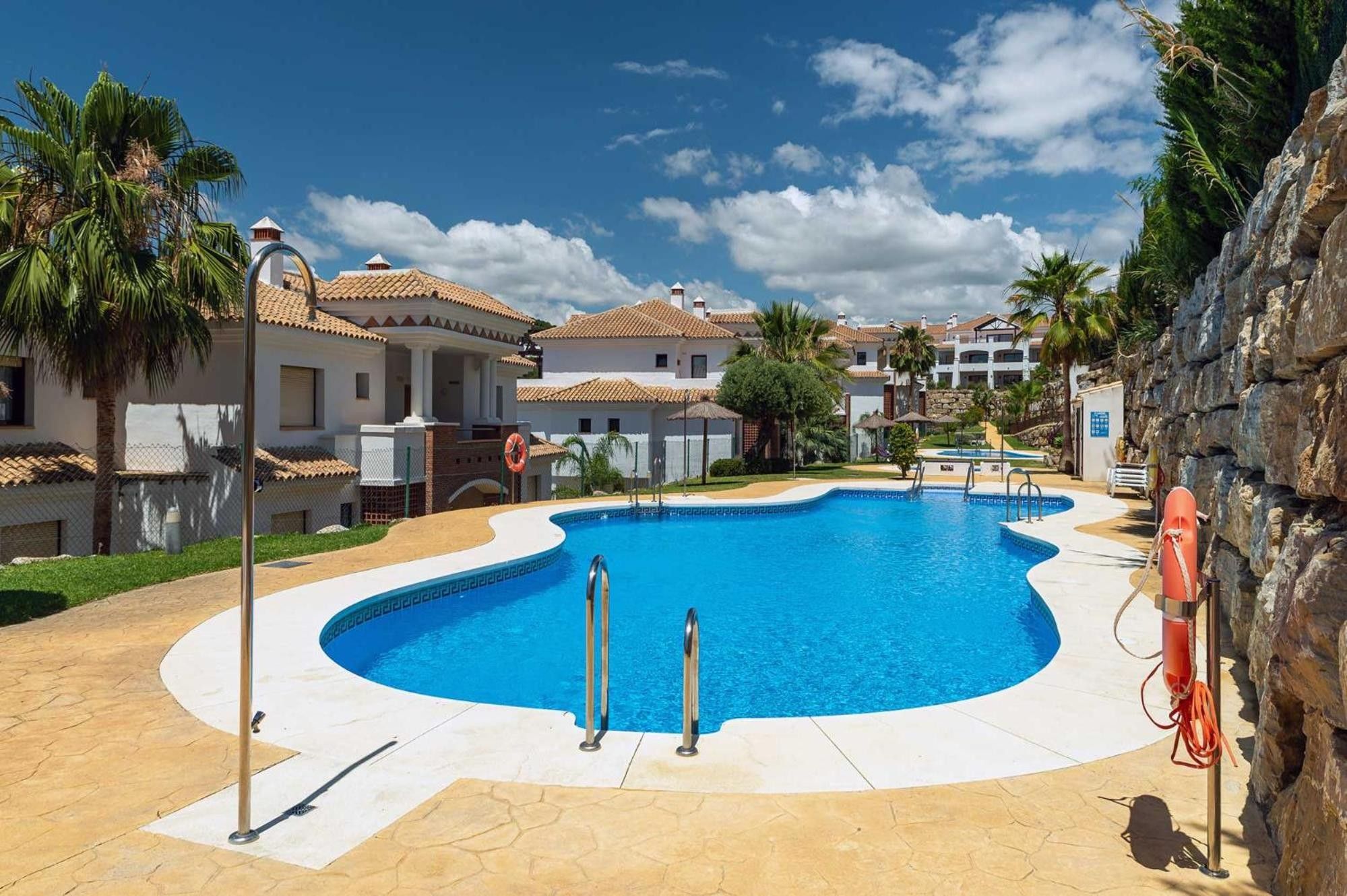 A swimming pool with a curved edge, surrounded by a paved deck, palm trees, and buildings with tiled roofs.