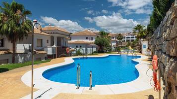 A swimming pool with a curved edge, surrounded by a paved deck, palm trees, and buildings with tiled roofs.