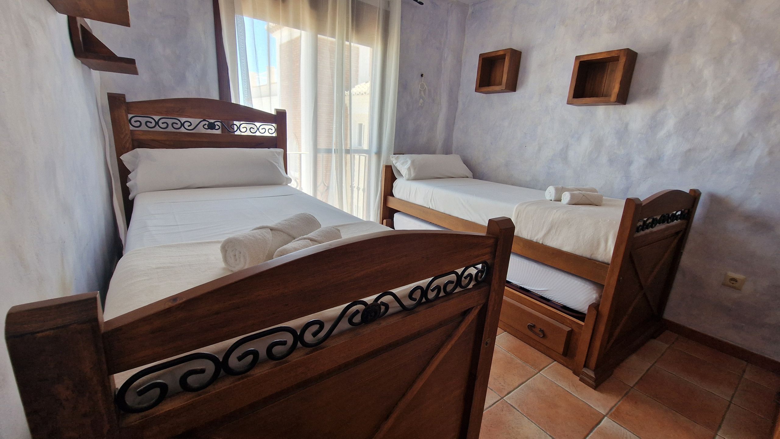 A room with two wooden beds, a window with curtains, and a wall-mounted shelf.