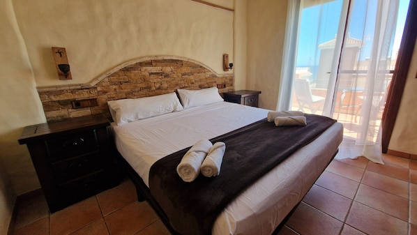 Habitación de hotel con cabecera de piedra, cama con ropa de cama blanca, manta marrón oscuro y un juego de toallas.