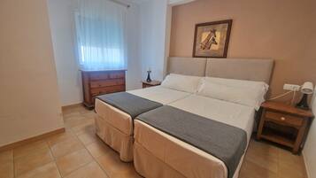 Una cama doble con cabecera, una mesita de noche con lámpara, un aparador de madera y una ventana con cortinas translúcidas.