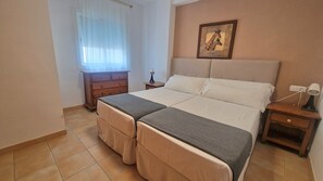 Una cama doble con cabecera, una mesita de noche con lámpara, un aparador de madera y una ventana con cortinas translúcidas.