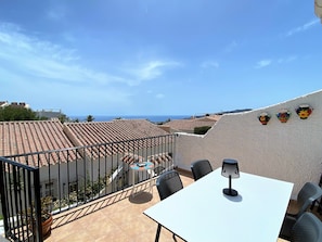 Eine Terrasse mit weißem Tisch und Stühlen, einem schwarzen Metallgeländer und Blick auf das Meer.