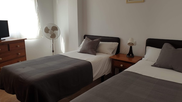 Habitación de hotel con dos camas, una mesita de noche con lámpara, un aparador y un ventilador de pie.