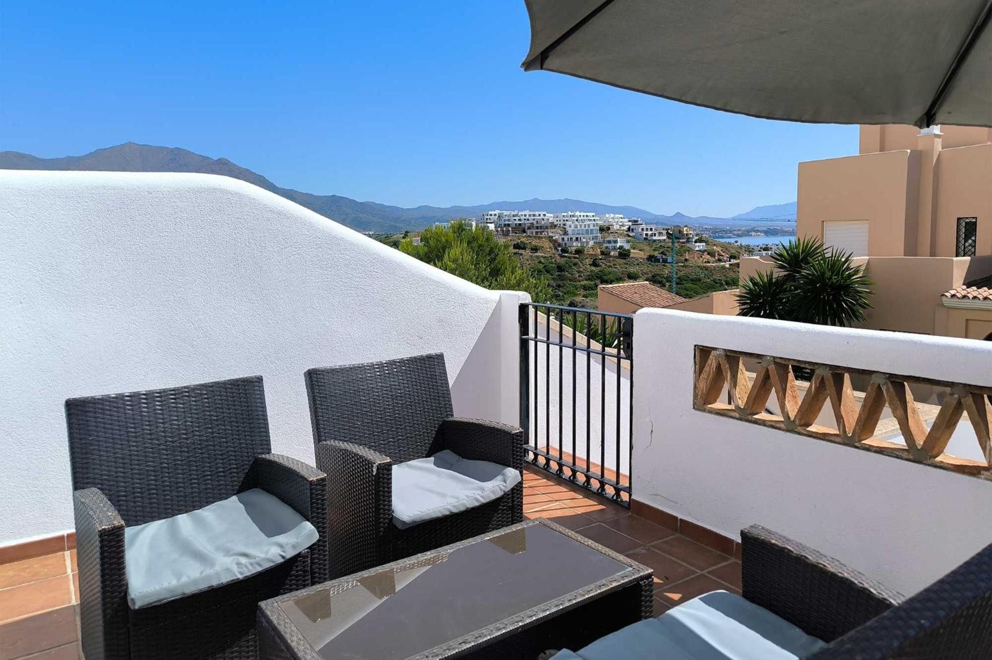 A terrace with wicker furniture and a glass table, overlooking a coastal town and mountains.