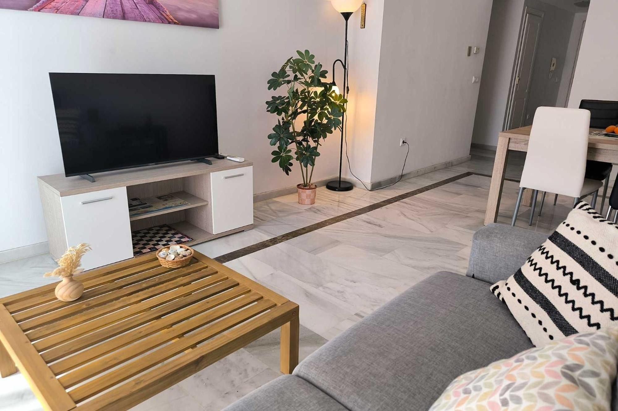 Una sala de estar con un televisor de pantalla plana, una mesa de centro de madera, un sofá gris, una planta en maceta y una lámpara de pie.
