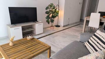 Una sala de estar con un televisor de pantalla plana, una mesa de centro de madera, un sofá gris, una planta en maceta y una lámpara de pie.