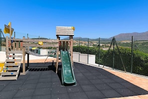 Un parque infantil con un tobogán y estructuras para escalar, rodeado por una valla y con vistas a un paisaje montañoso.