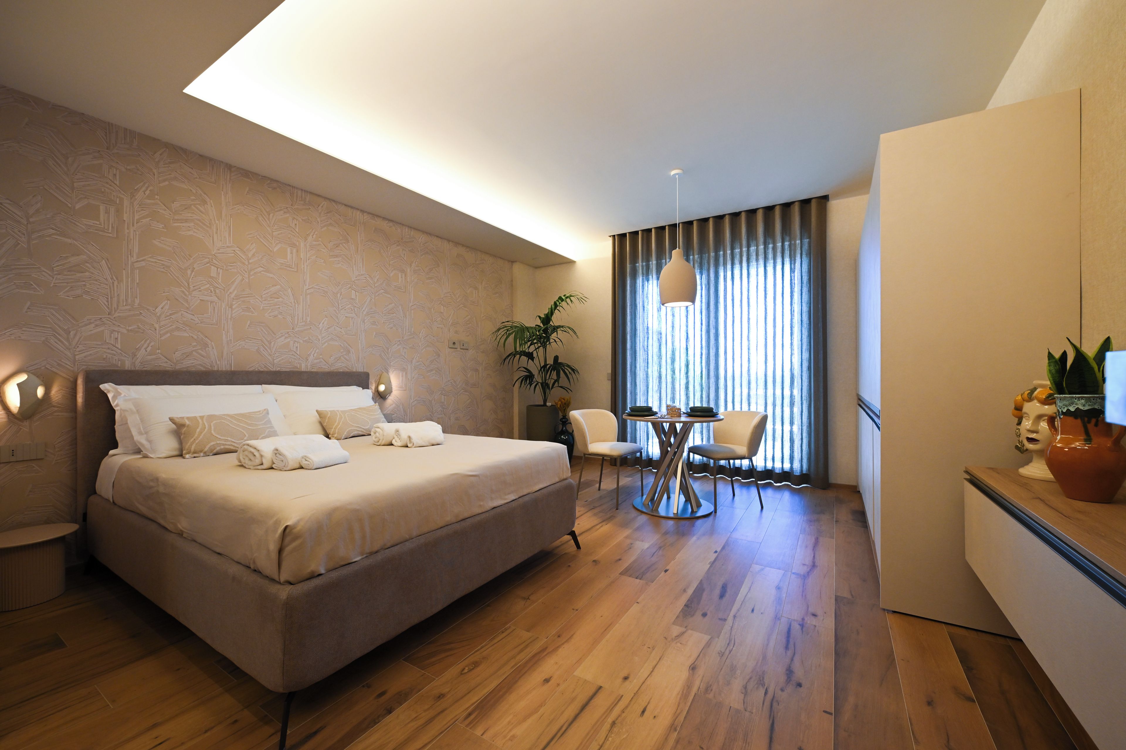 A hotel room with a bed, bedside table, lamp, curtains, and a dining area with chairs and a table.