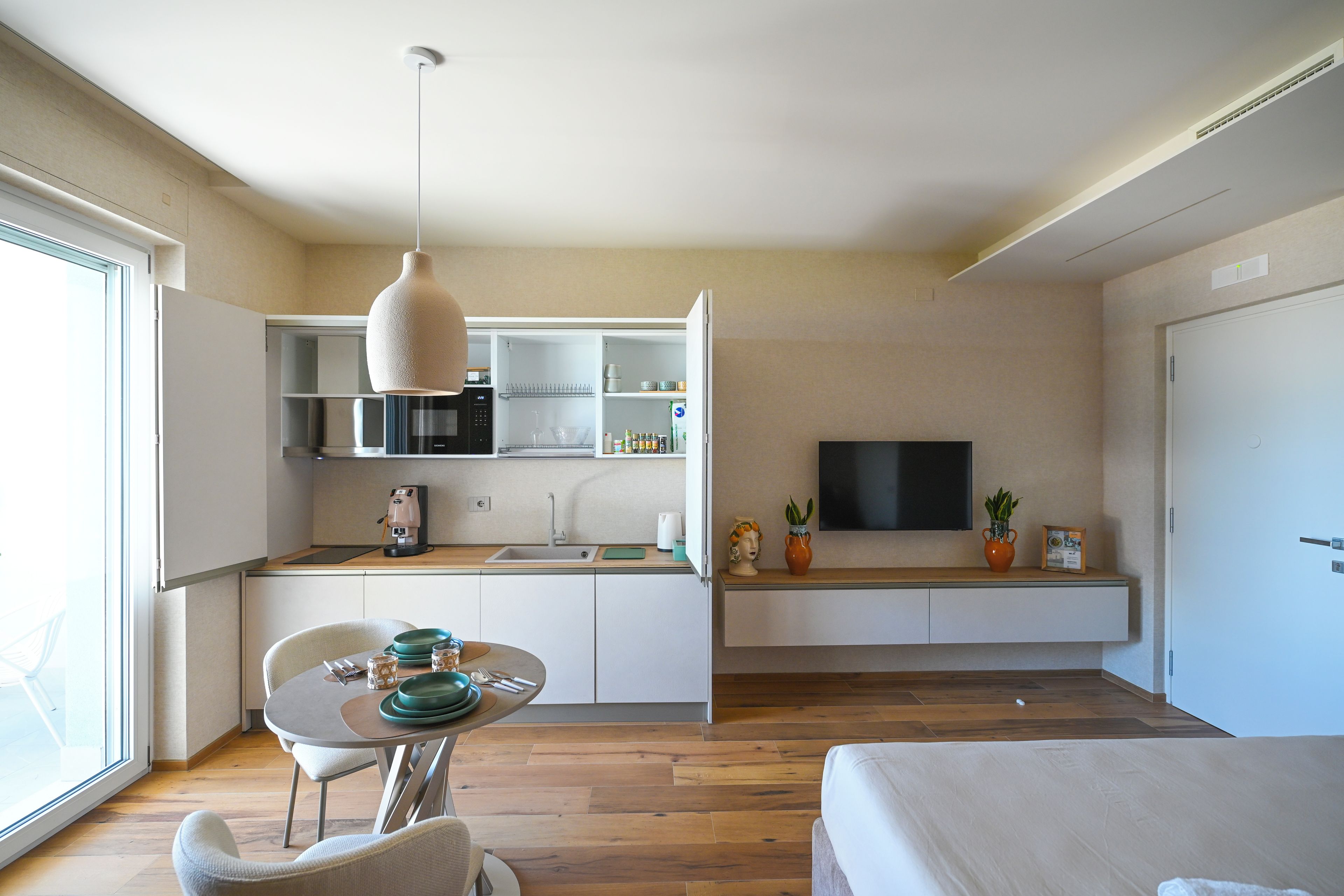 A modern kitchen with a dining area, a TV mounted on the wall, and a large window providing natural light.