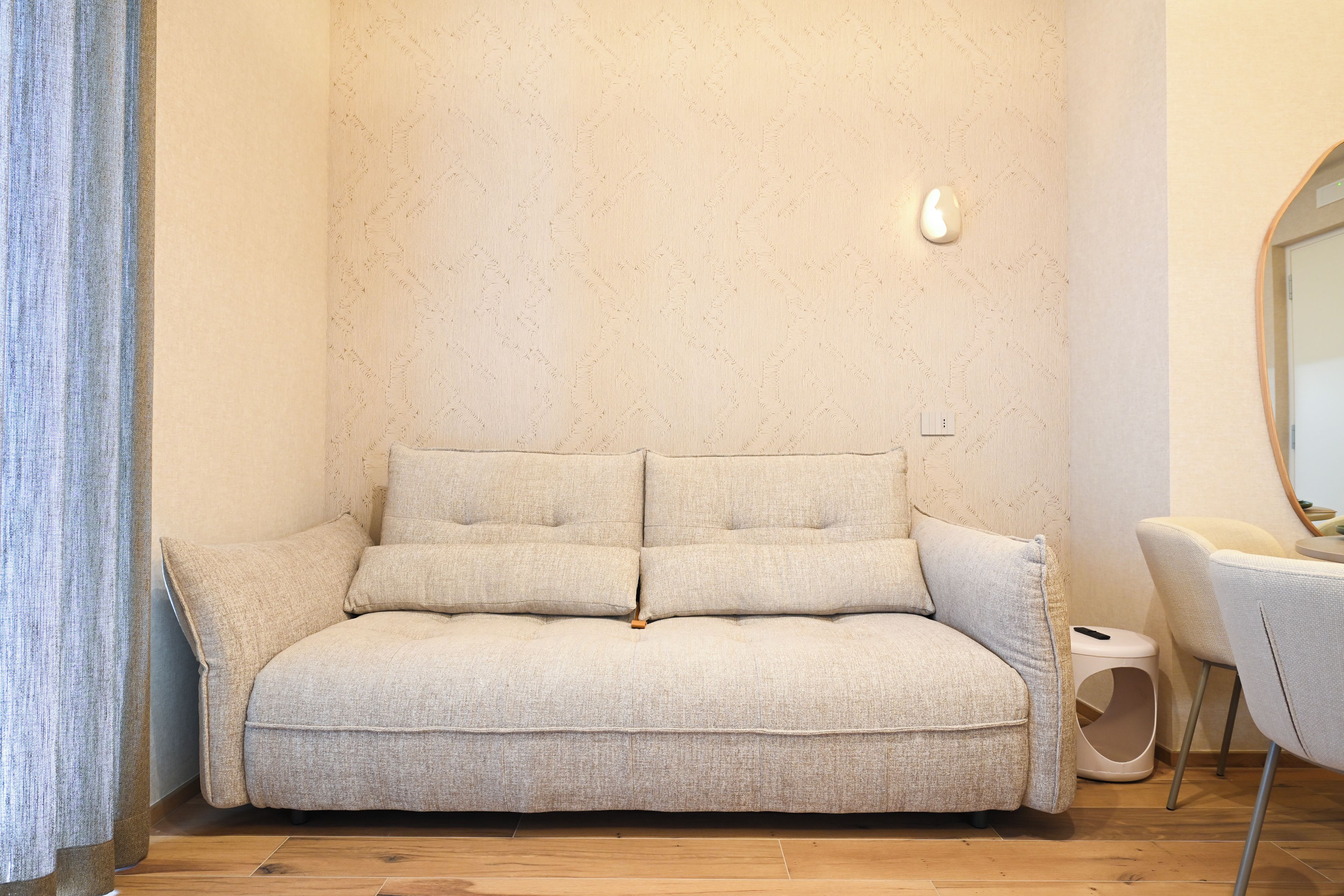 Un canapé beige, recouvert d’un tissu texturé, est placé contre un mur orné d’un motif discret.