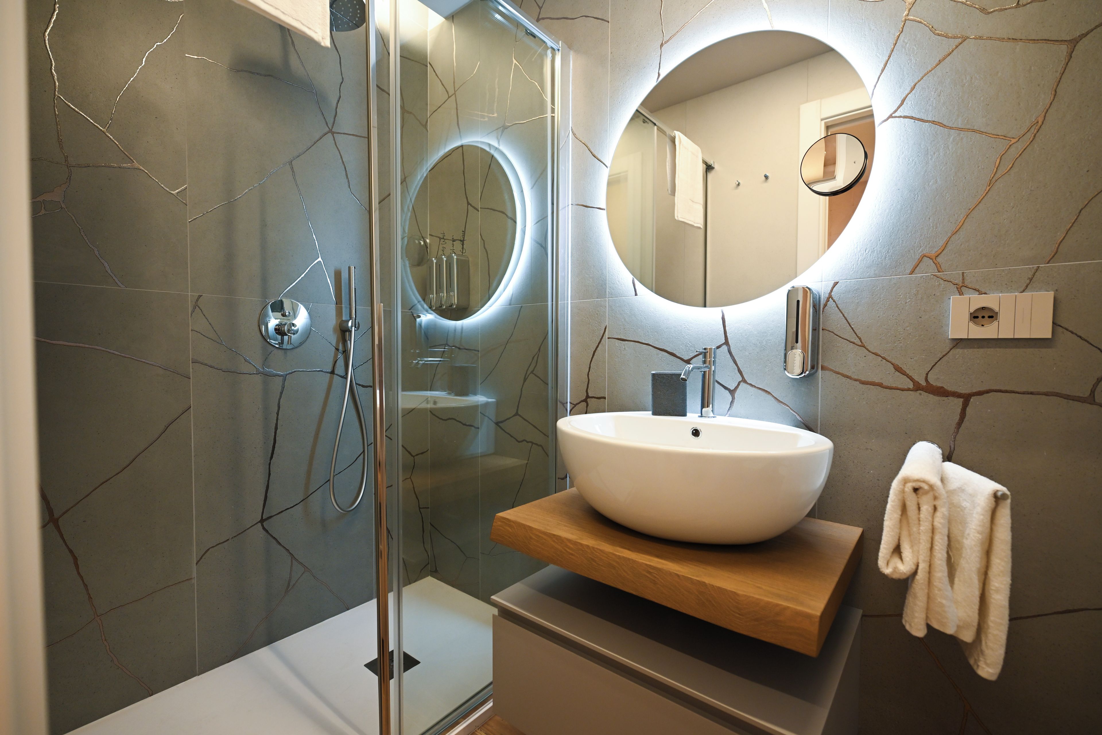 Un baño moderno con ducha envuelta en vidrio, un espejo redondo y un lavamanos blanco sobre una encimera de madera.