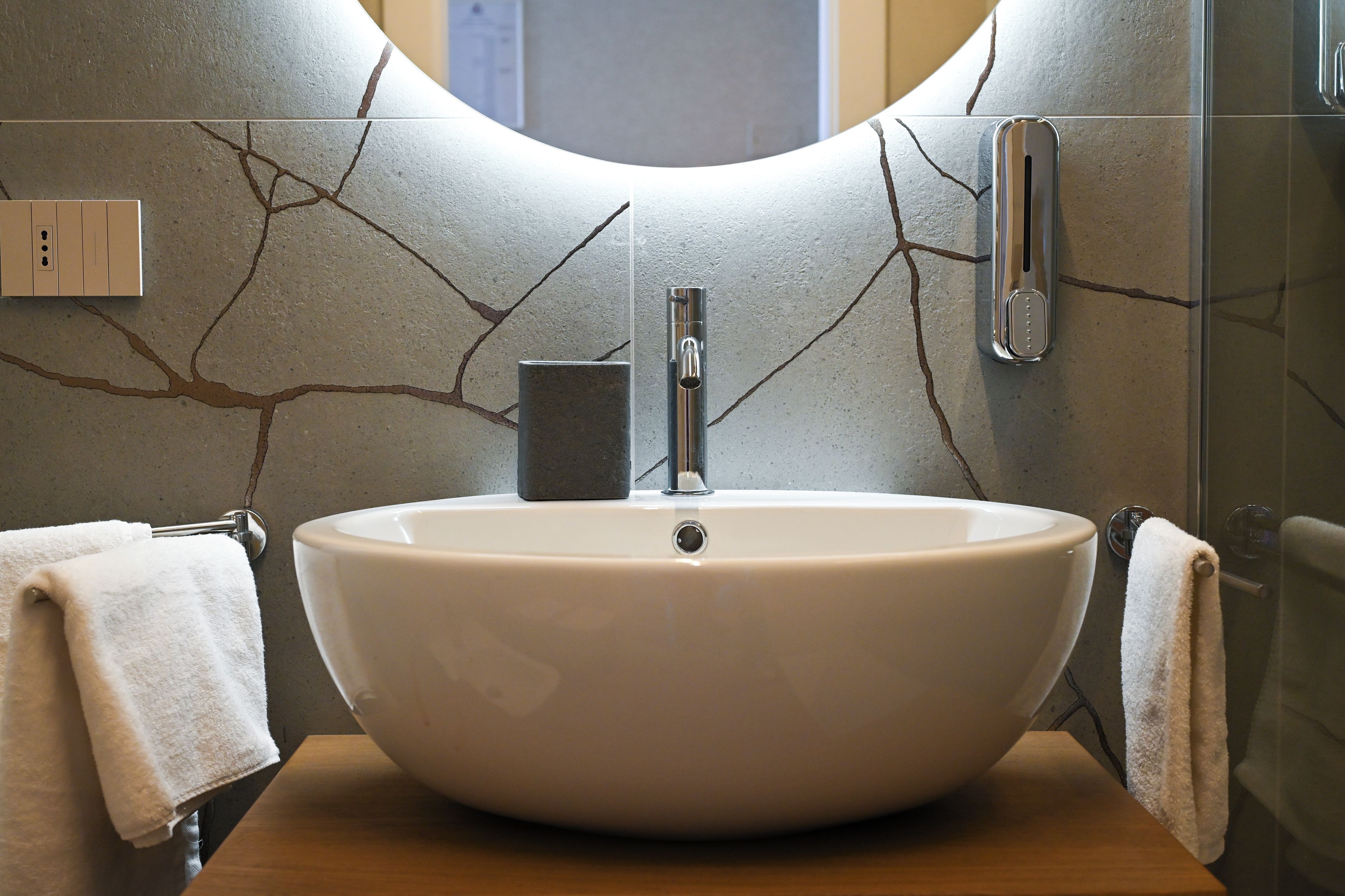 A bathroom with a large white vessel sink, a faucet, and a soap dispenser. There are towels on a rack and a mirror above the sink.