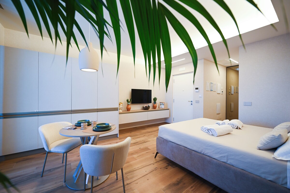 A modern hotel room with a bed, a dining table with chairs, a TV, and a large plant.