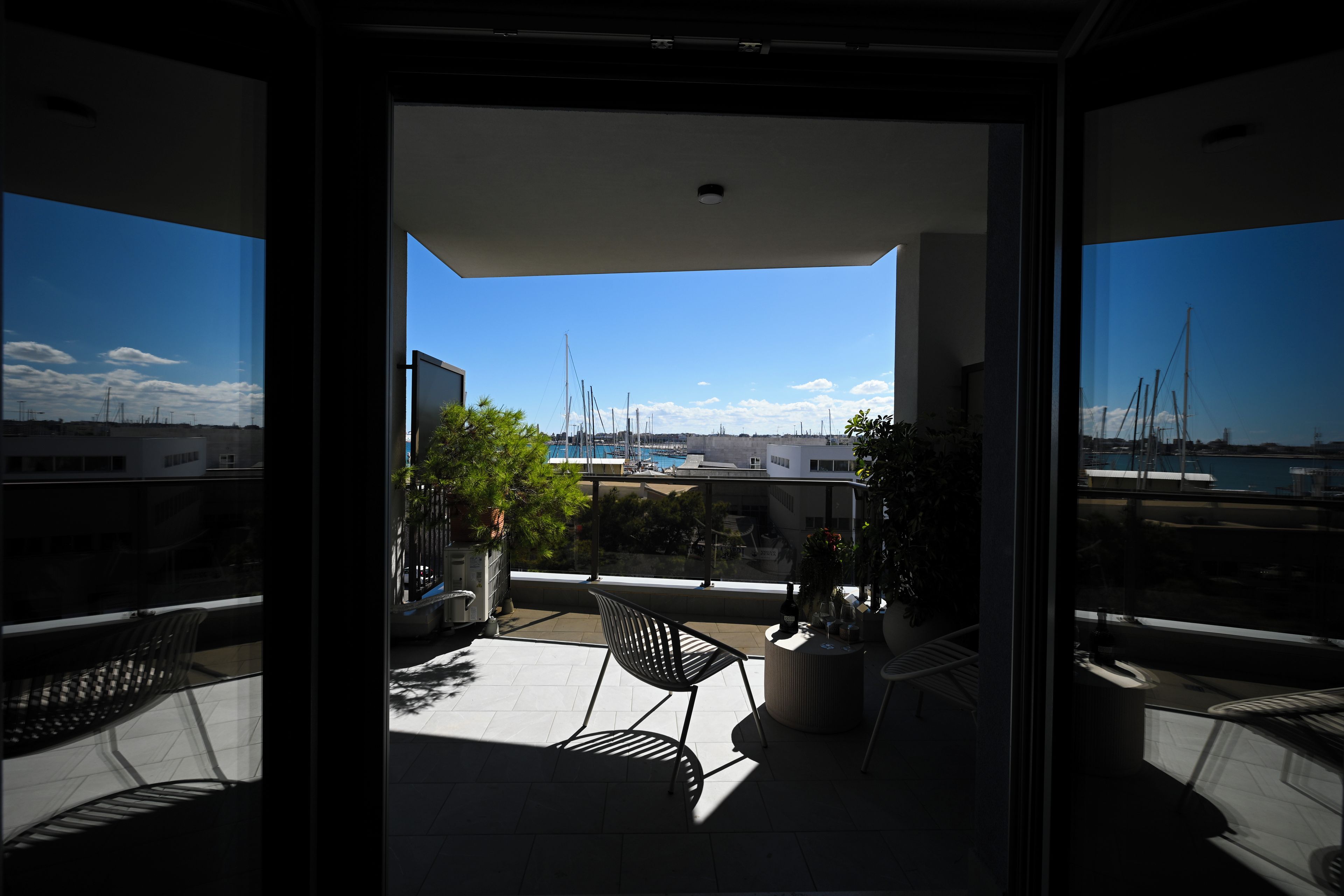 A balcony with a view of the sea, a chair, a table with a bottle and glasses, and potted plants.