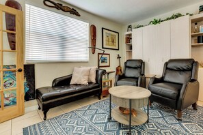 Una sala de estar con un sofá de cuero negro, dos sillones de cuero negro, una mesa de centro redonda, un tapete estampado y una ventana con persianas.