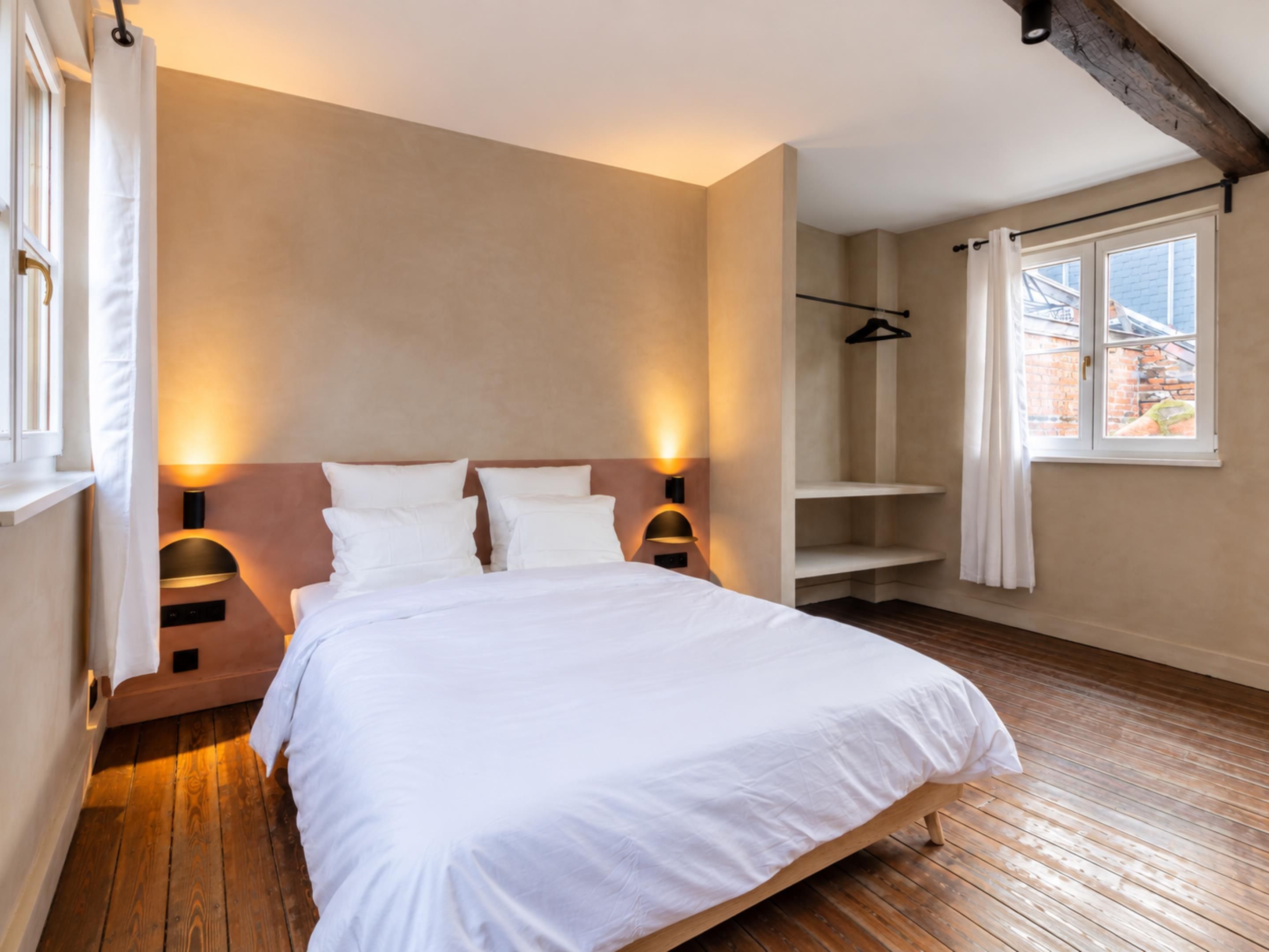 Ein Schlafzimmer mit Holzboden, einem Bett mit weißer Bettwäsche und einem Fenster mit Blick auf Gebäude.