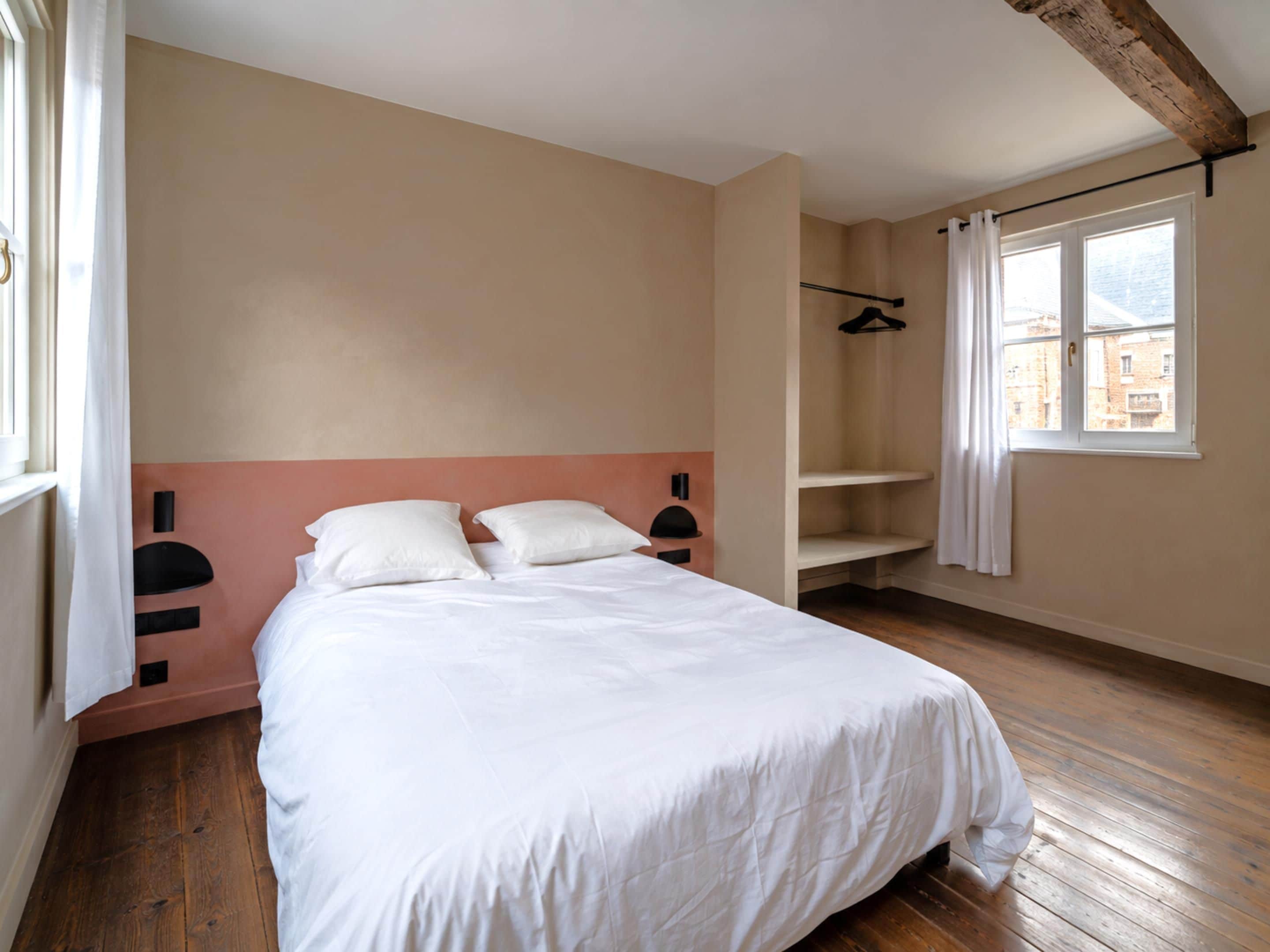 A bedroom with a bed, a window, a coat rack, and a wooden floor.