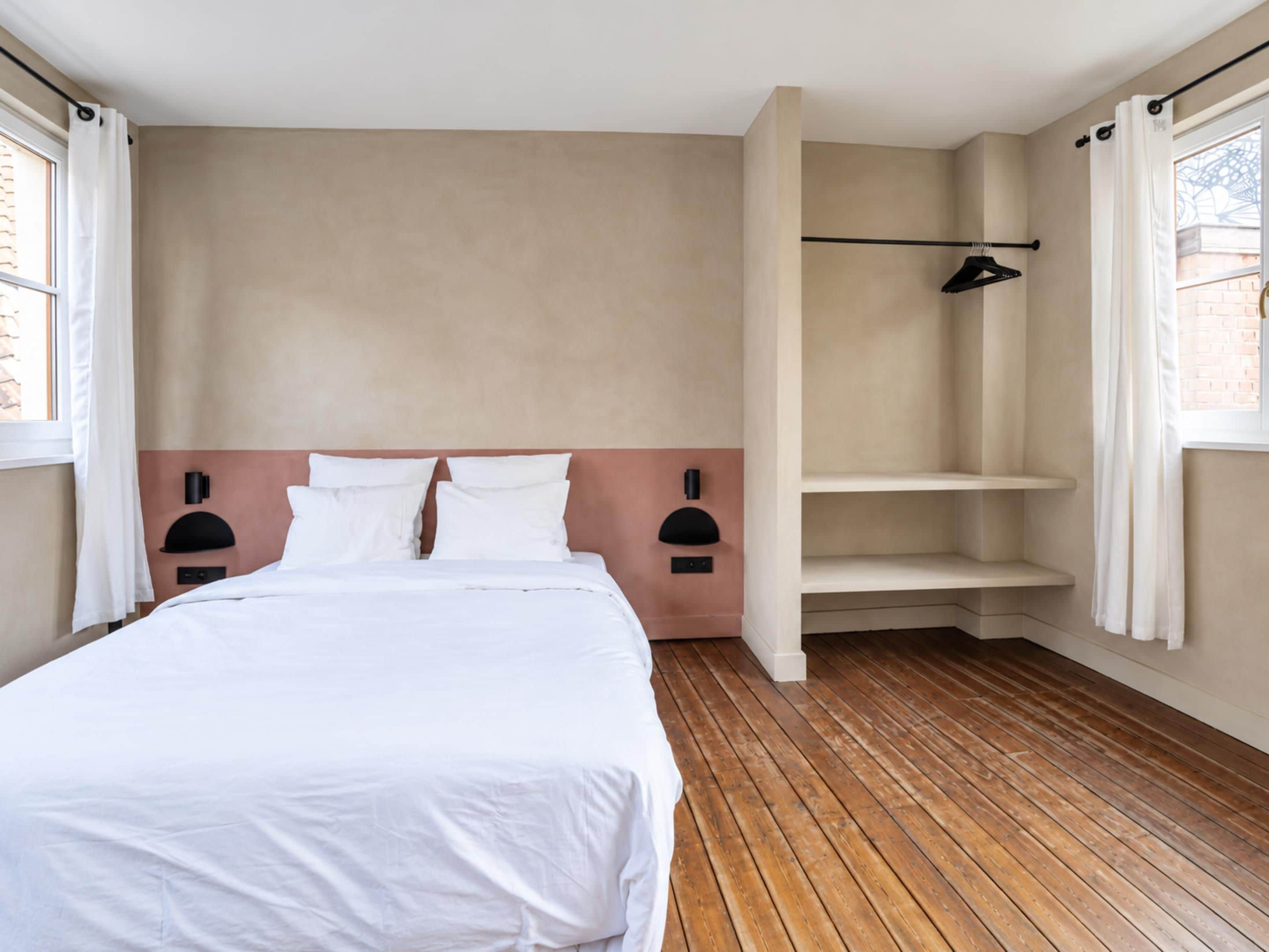 Een slaapkamer met een houten vloer, een bed met wit beddengoed, een wandplank en een raam met gordijnen.