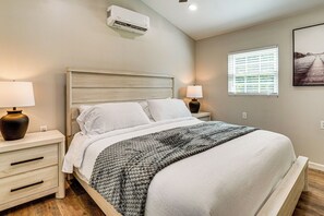 A bedroom with a bed, bedside tables, a lamp, an air conditioner, a window with blinds, and a framed picture on the wall. - New To Market Riverfront Pigeon Forge Retreat (Pigeon Forge)