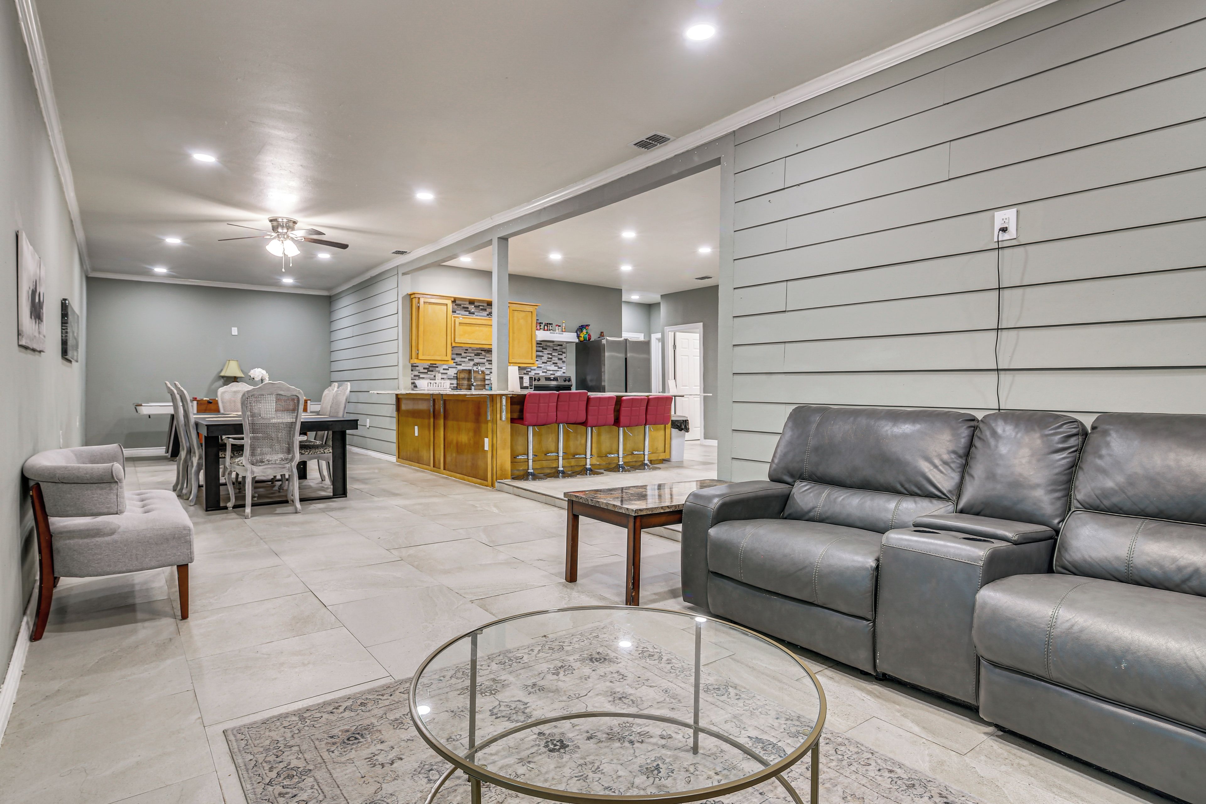 A modern living room with a grey leather sofa, a glass coffee table, and a dining area with a kitchen in the background.