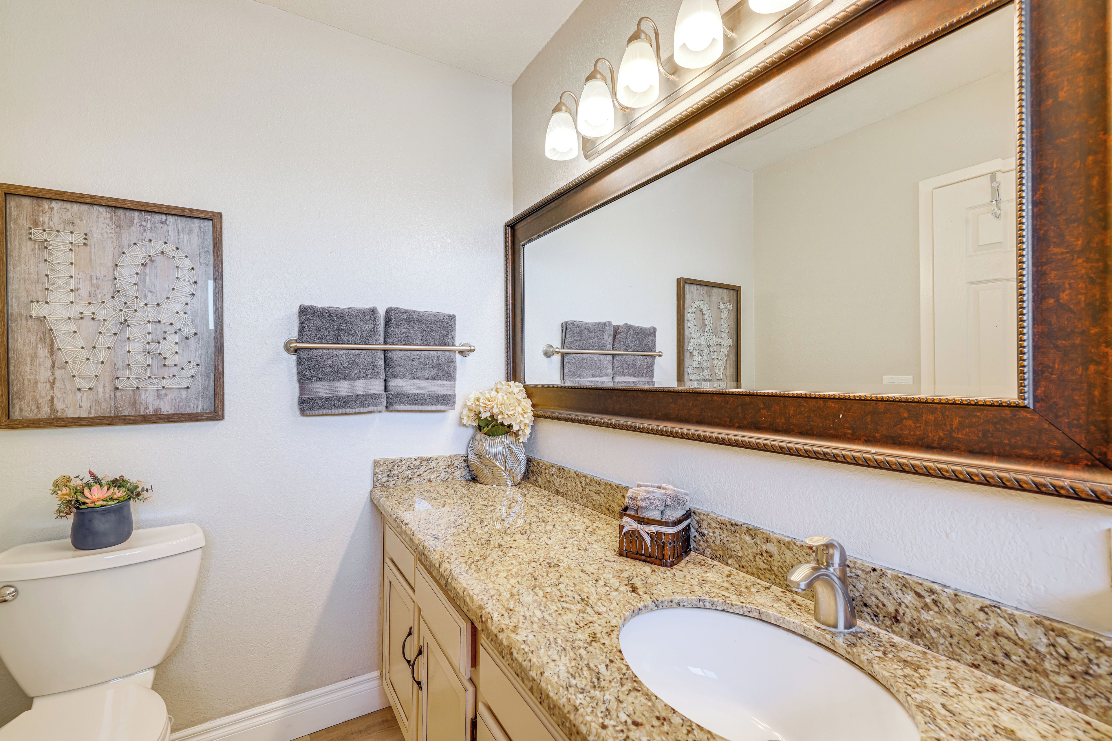 A bathroom with a toilet, sink, vanity, mirror, towel rack, and decorative wall art.