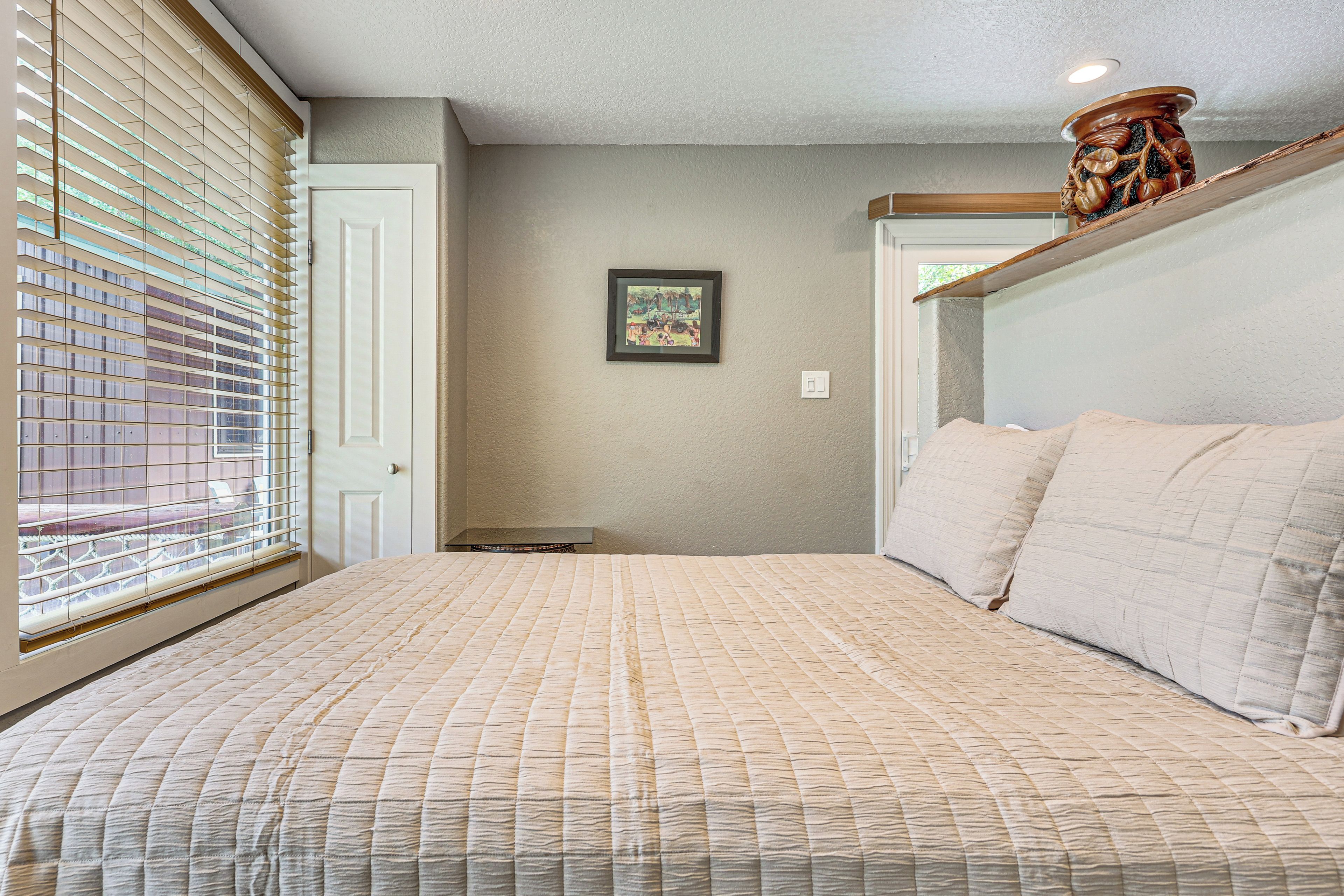 A bedroom with a bed, a window with blinds, a door, and a framed picture on the wall.