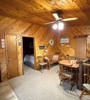 Ein gemütliches Blockhaus-Interieur mit Holzbalkendecke, einem Esstisch mit Stühlen, einem Bett und einer kleinen Küchenzeile.