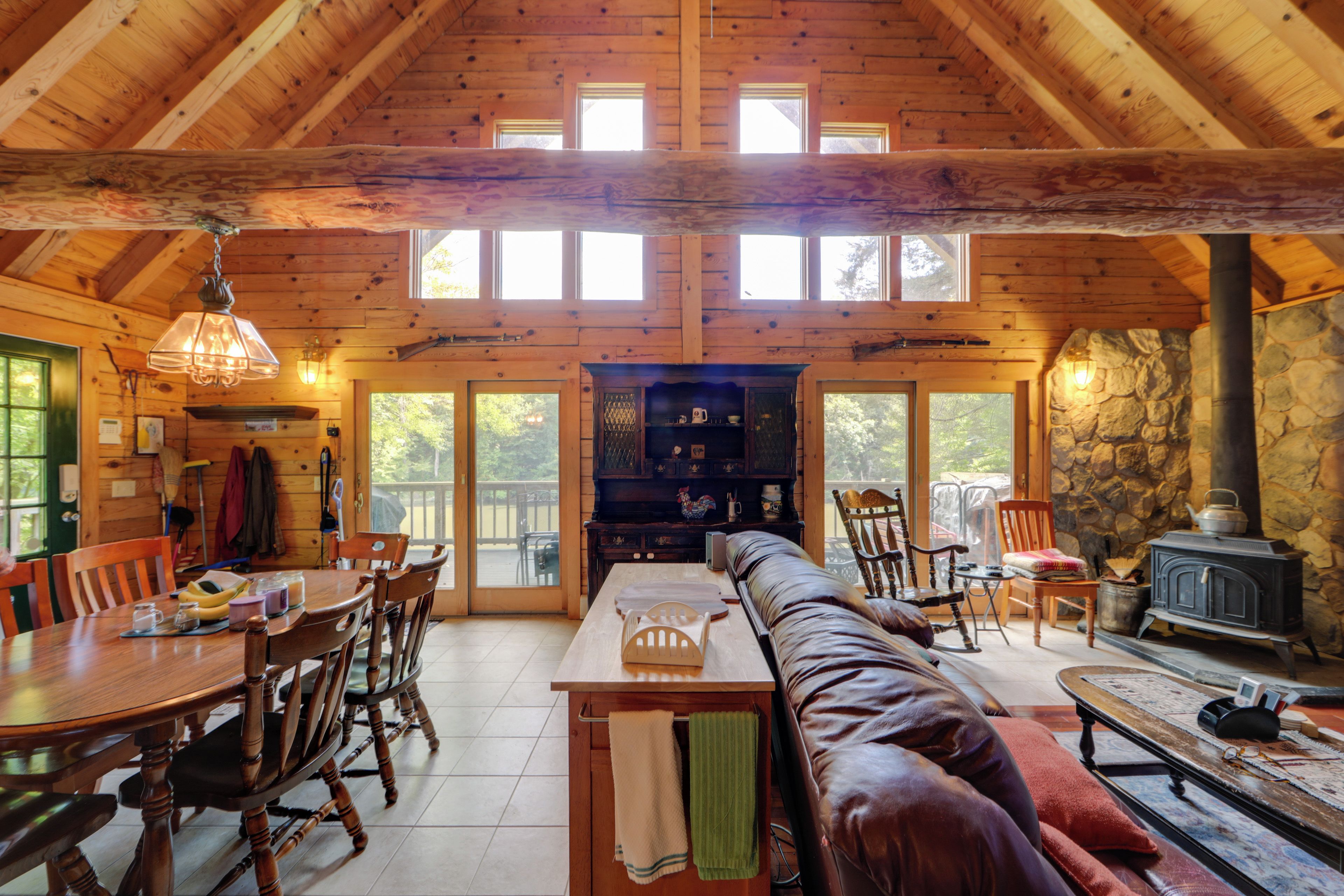 A spacious cabin interior with a high ceiling, wooden walls, a stone fireplace, and a dining area with a long table and chairs.