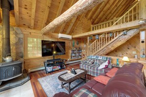 Interior acolhedor de uma cabana de madeira com lareira, televisão, estantes de livros e uma escada.