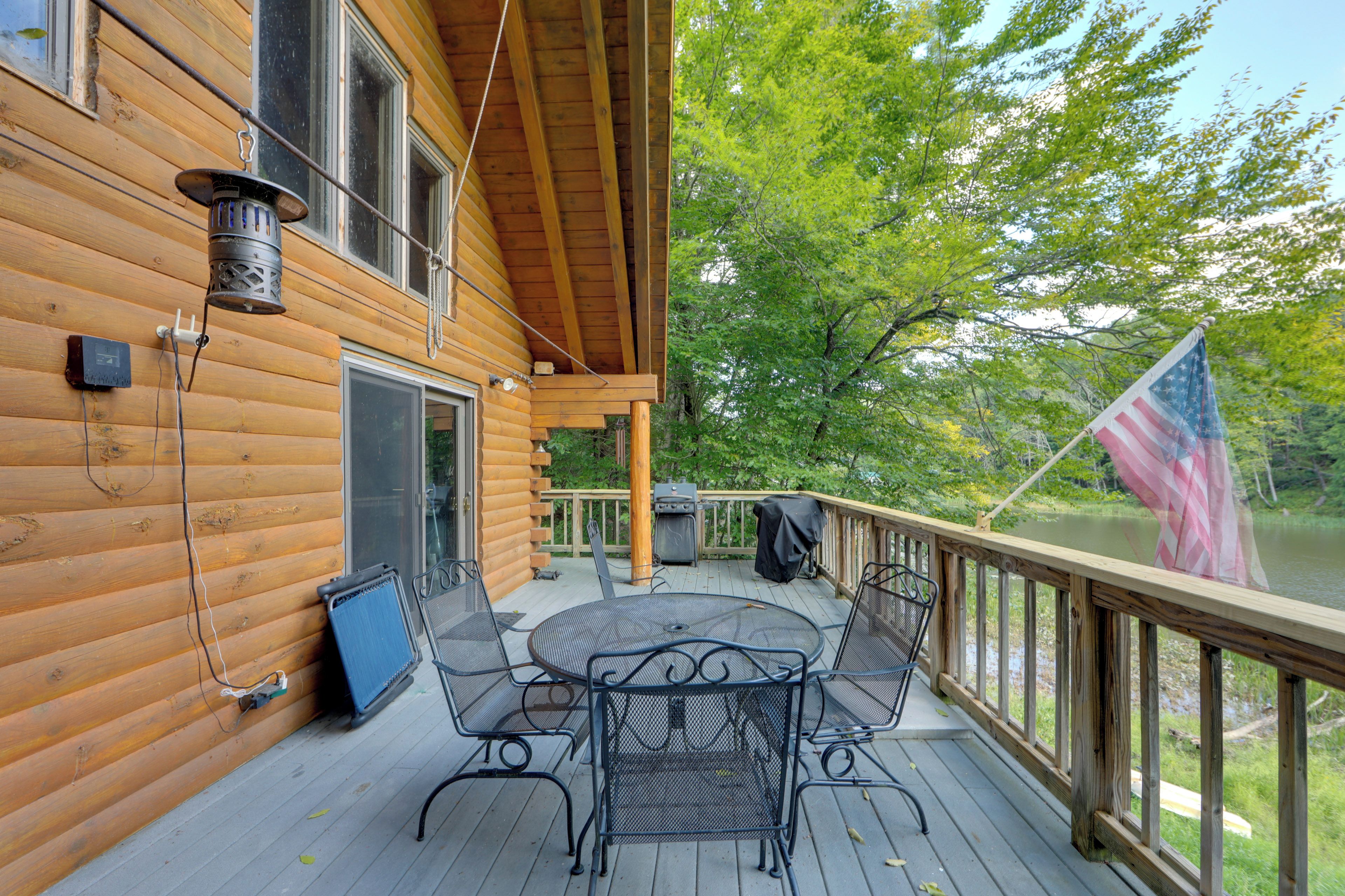 A wooden deck with a table and chairs, a flag, and a view of trees and water.