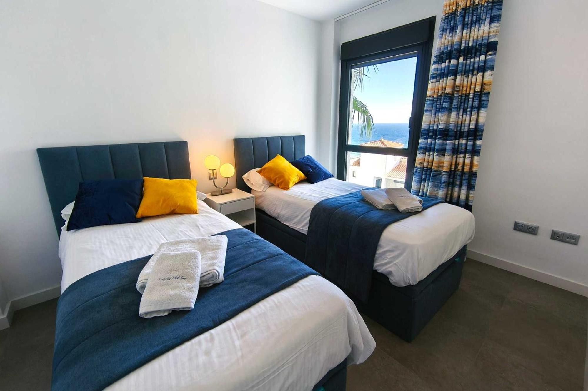 Habitación de hotel con dos camas, una mesita de noche con lámpara y una ventana con cortinas.