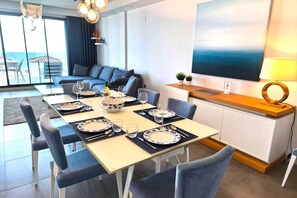 Un comedor con mesa dispuesta para seis personas, un sofá, un televisor y vistas al mar.