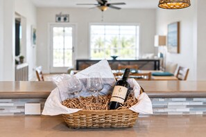 A kitchen area with a dining table, chairs, and a basket containing a bottle of wine and two glasses on the counter. - Beachfront Access | 4BR | Hot Tub | Ocean Views (Galveston)