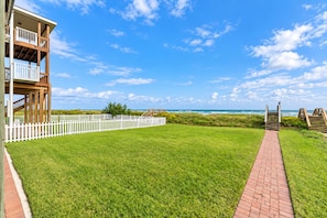A grassy area with a white fence, a brick pathway, and a wooden structure with balconies. - Beachfront Access | 4BR | Hot Tub | Ocean Views (Galveston)