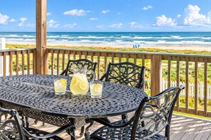A round metal table with chairs on a wooden deck overlooking a beach. - Beachfront Access | 4BR | Hot Tub | Ocean Views (Galveston)