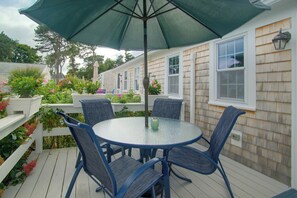 A covered outdoor seating area with a round table and four chairs, surrounded by a white fence and various plants. - Walk to Beach & Eats! Dennis Port Cottage-2 Decks (Dennis Port)
