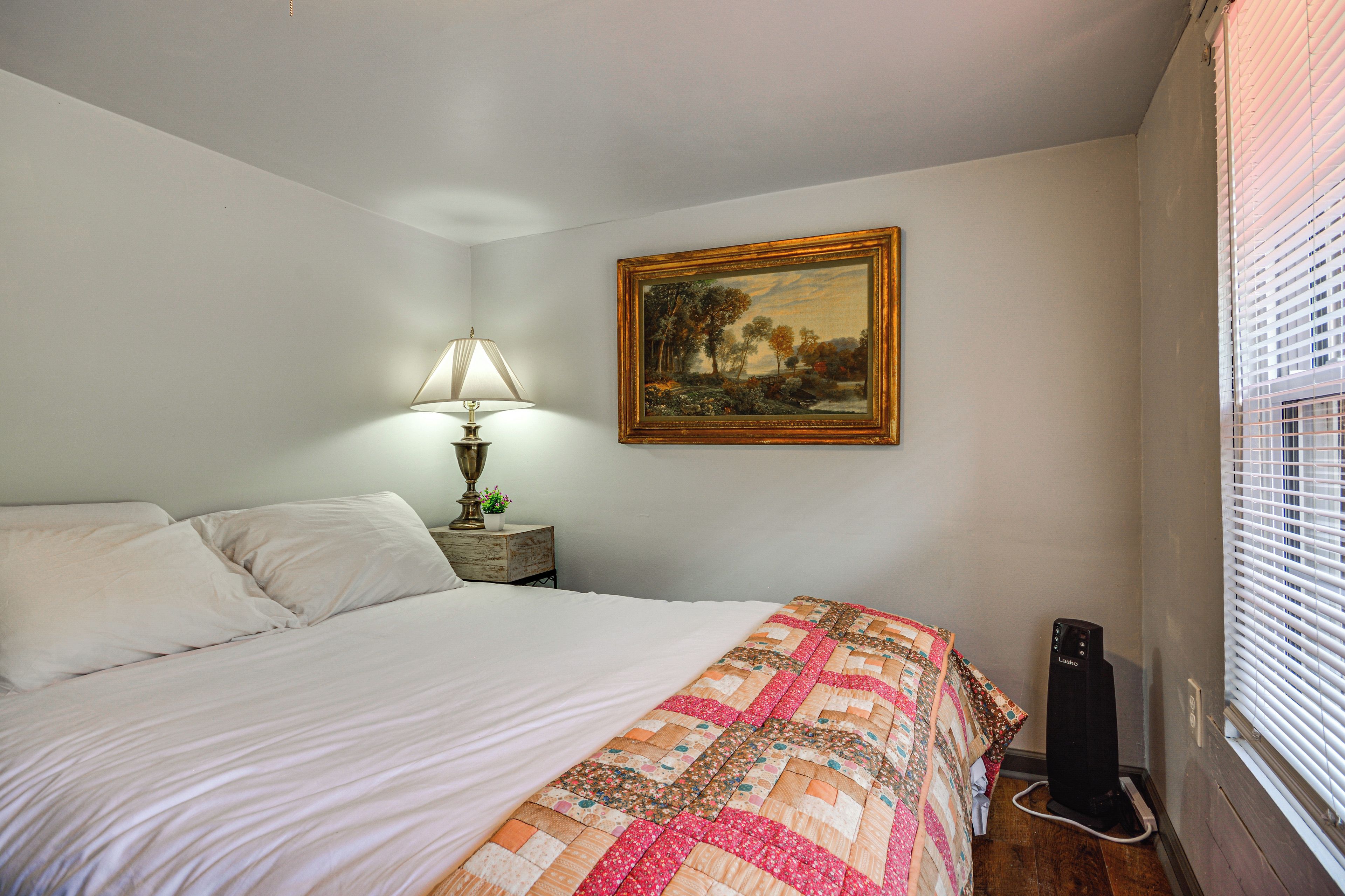 A bedroom with a bed, a nightstand, a lamp, a painting, and a window with blinds.