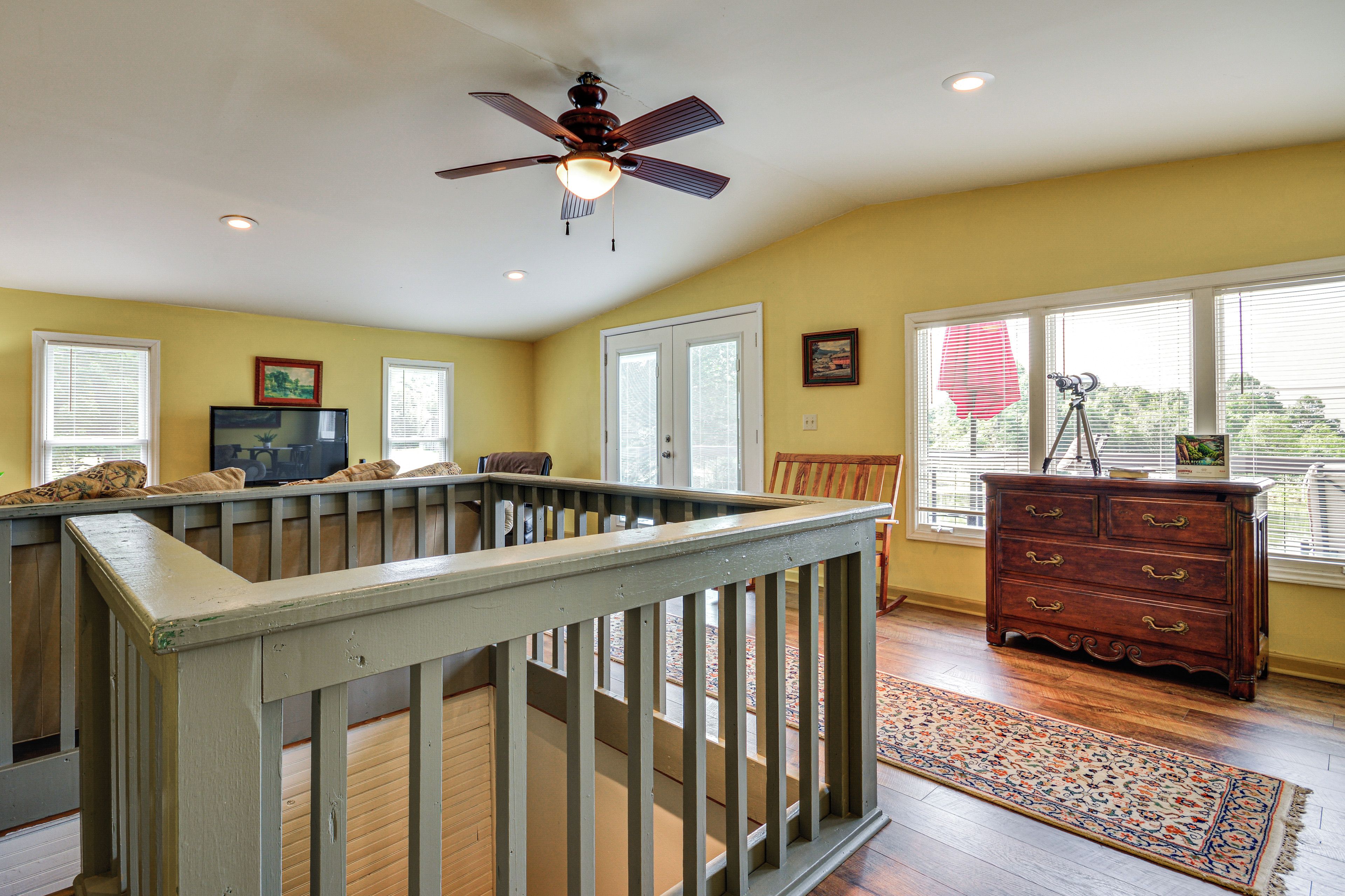 A room with a wooden railing, a ceiling fan, a dresser, and a window with blinds.