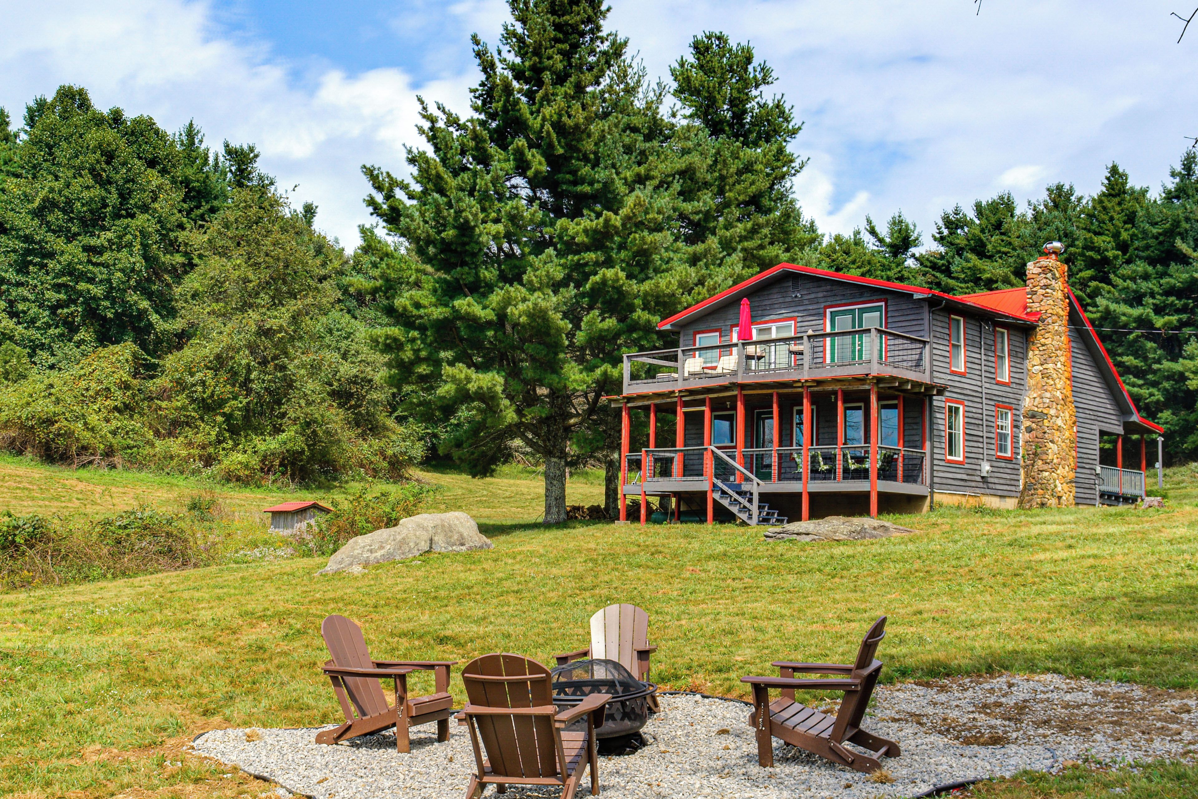 A two-story cabin with a red roof, a stone chimney, and a deck with chairs.