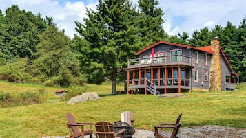A two-story cabin with a red roof, a stone chimney, and a deck with chairs.