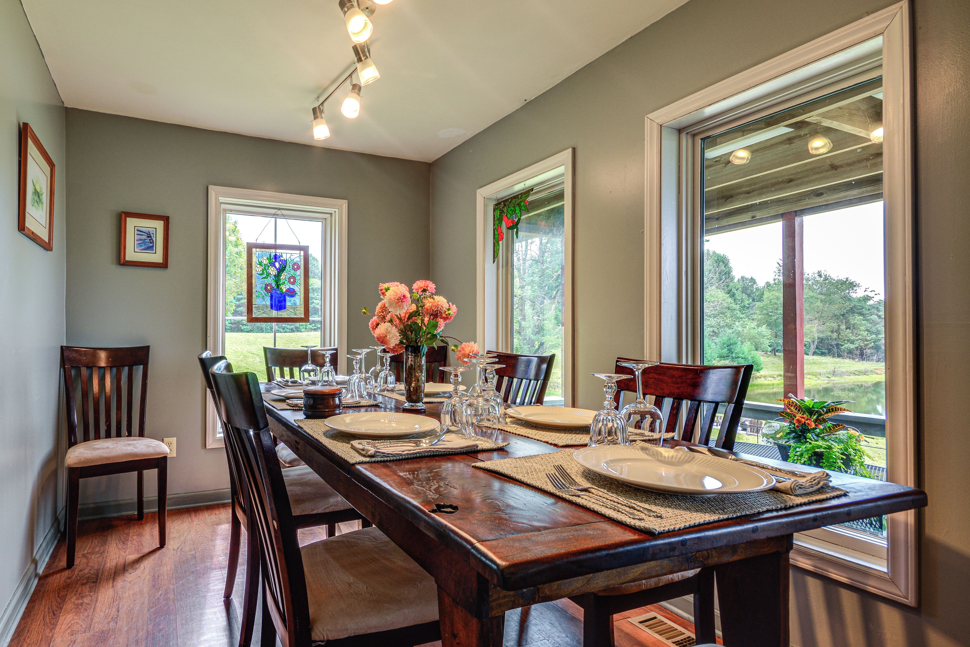 A dining room with a long wooden table set for a meal, flanked by chairs, with large windows offering a view of the outdoors.