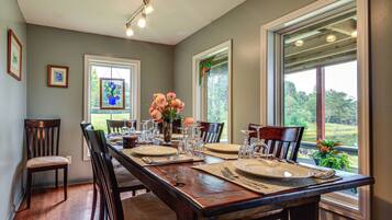 A dining room with a long wooden table set for a meal, flanked by chairs, with large windows offering a view of the outdoors.