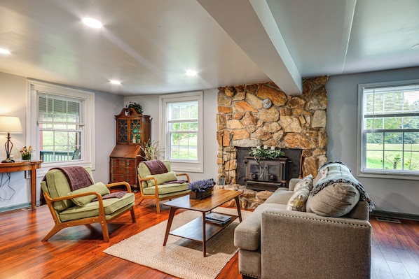 A living room with a stone fireplace, wooden furniture, and large windows. - On 62-acre Farm! Waterfront Home w/ Decks & Views (Meadows Of Dan)