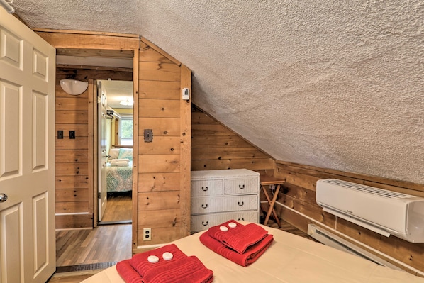 A room with a sloped ceiling, wooden walls, a bed with red towels, a white dresser, and an air conditioning unit.