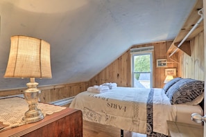 Un dormitorio con techo inclinado, paredes de madera, una cama con ropa blanca, una lámpara en una mesita de noche de madera y una ventana con vistas a follaje.