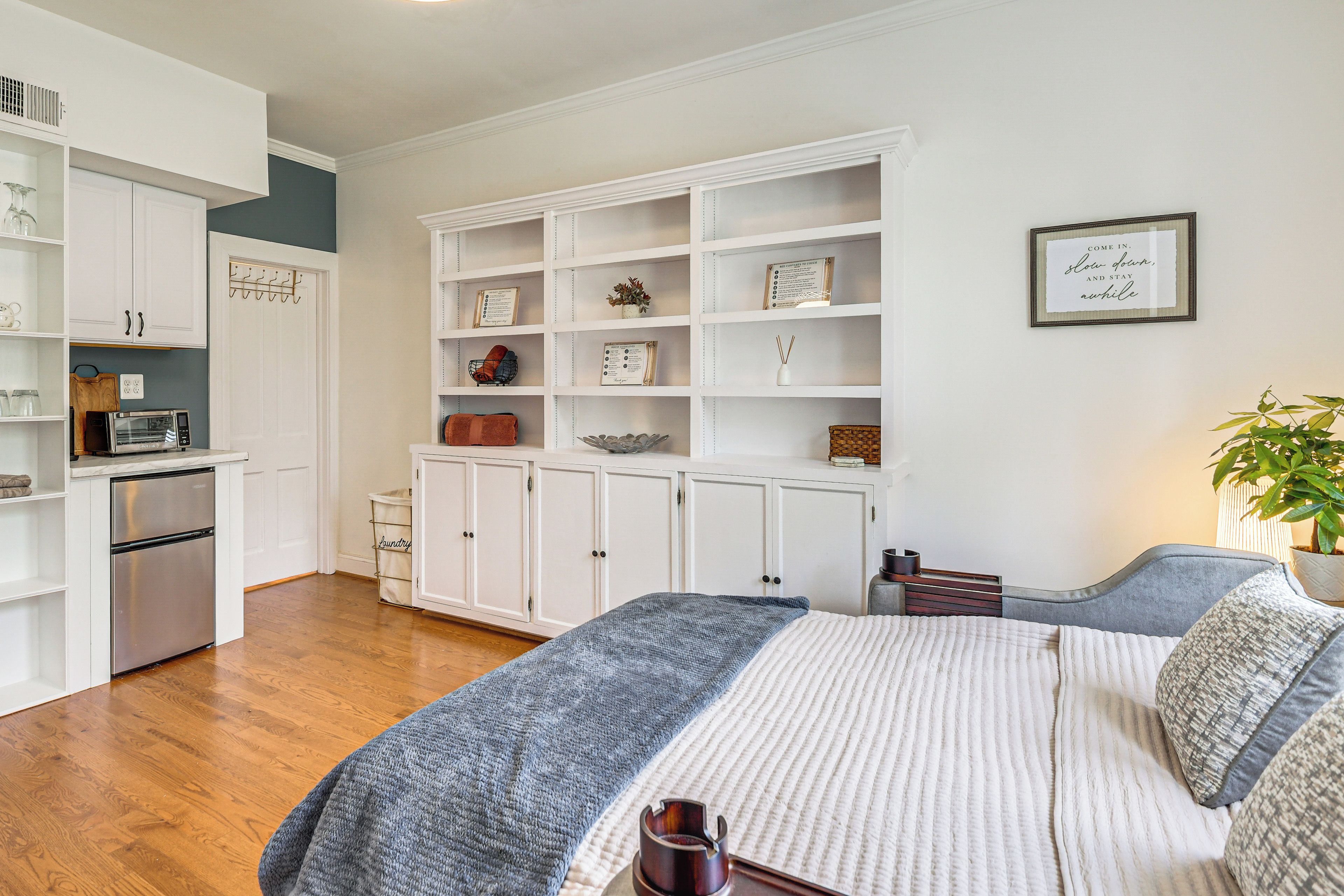 A bedroom with a bed, a bedside table, a nightstand, a wall-mounted shelf with decorative items, and a potted plant.
